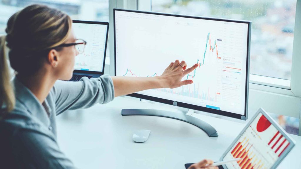 A woman is sitting at a white desk in front of a computer monitor and laptop. She is pointing to a chart on her monitor.